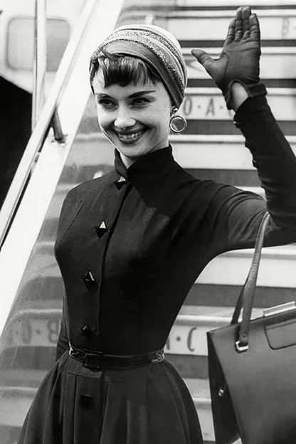 Black-and-white photograph of Audrey Hepburn waving from aircraft steps in a fitted dark dress, headscarf and gloves