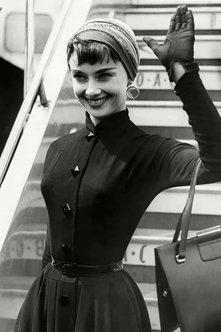 Black-and-white photograph of Audrey Hepburn waving from aircraft steps in a dark buttoned dress, headscarf and gloves