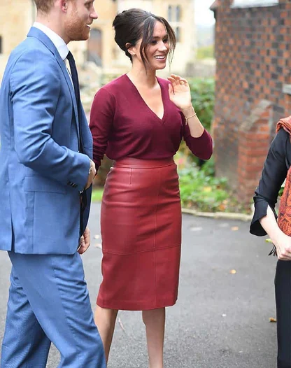 Meghan Markle in a burgundy knit top and red leather pencil skirt, smiling and talking with a man in a blue suit