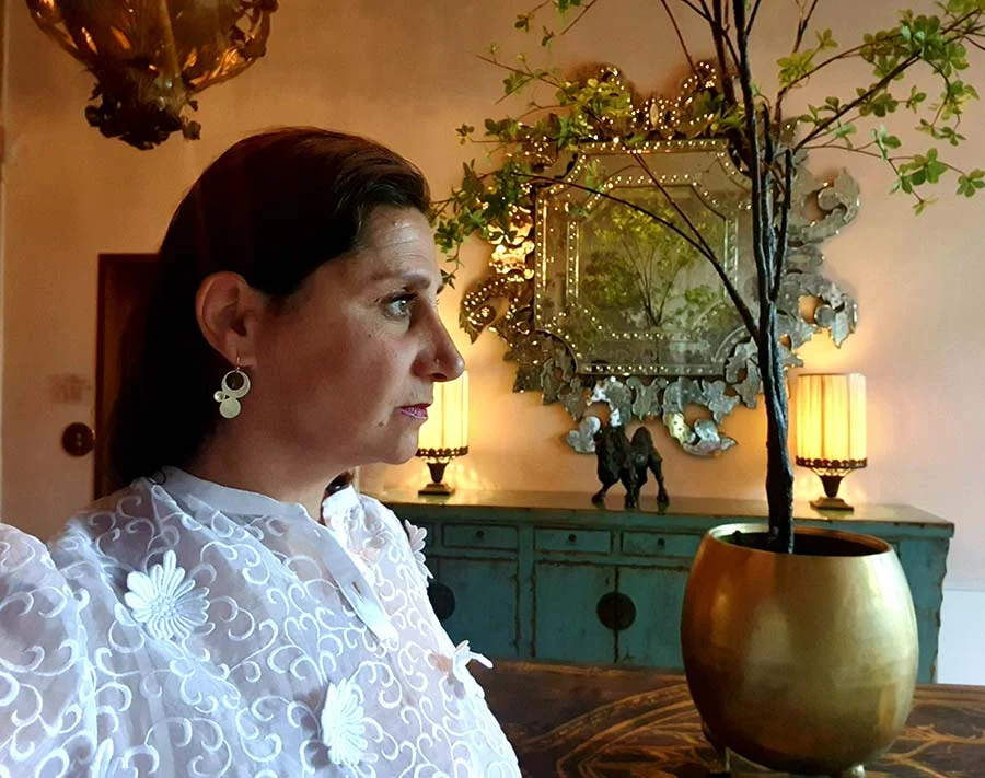 Gracie Opulanza in profile in a white lace blouse with flower appliqué, beside a gilt mirror and a gold bowl with a branch