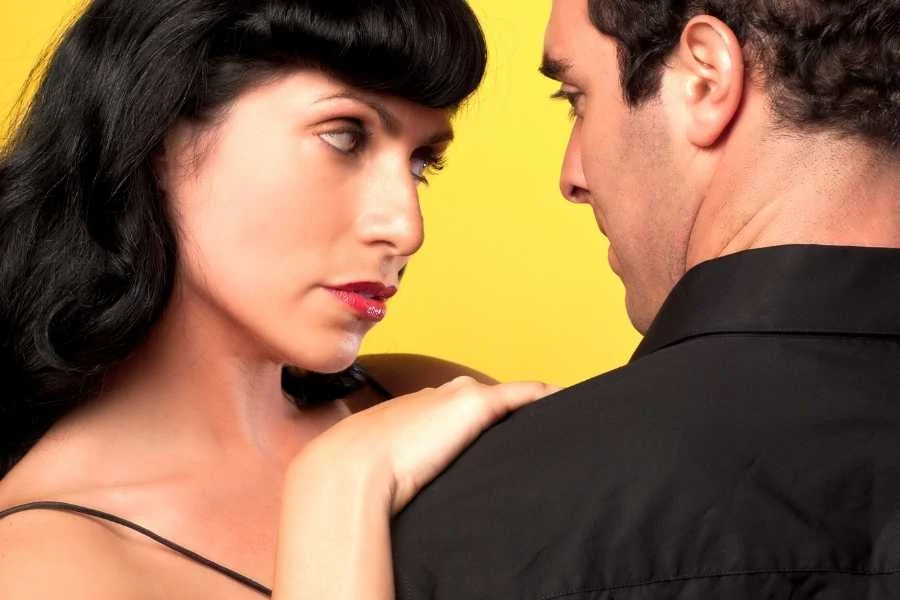 Woman with a blunt black Bettie Page fringe and red lipstick, leaning towards a man in a black shirt against a yellow wall