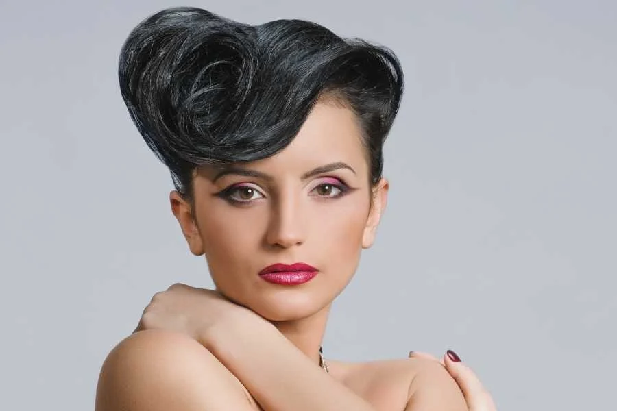Woman with a glossy black quiff swept up and over at the front, with winged eyeliner and berry lipstick