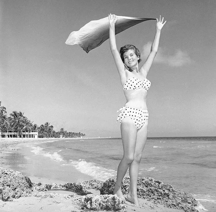 Black-and-white photograph of a smiling woman in a polka dot two-piece on a rocky beach, holding a cloth above her head