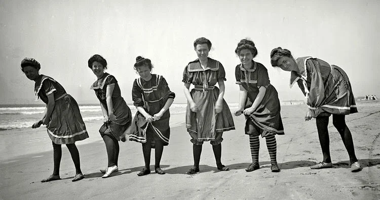 Black-and-white photograph of six women in dark Victorian bathing dresses with sailor collars, walking along a beach