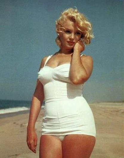 Marilyn Monroe on a beach in a white swimsuit with a sweetheart neckline, one hand touching her hair