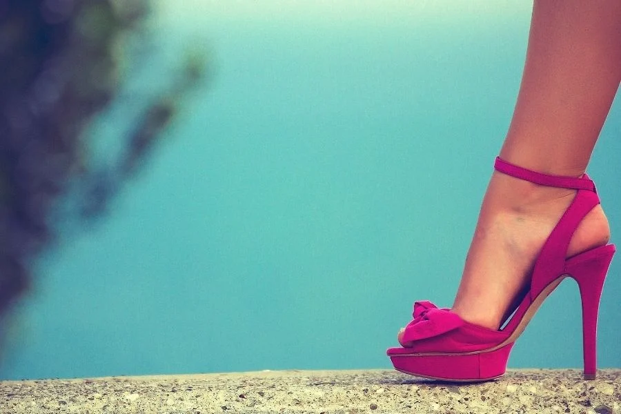 Pink suede platform sandal with a bow and ankle strap, worn on a stone ledge against a turquoise background