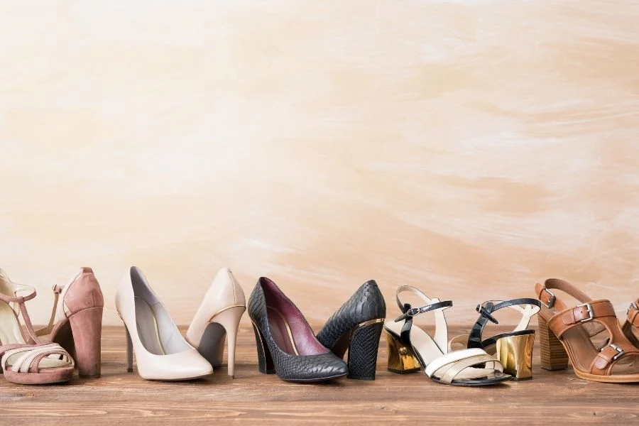 Row of women's shoes on a wooden floor: pink and cream pumps, black heels and sandals with gold heels, and tan sandals
