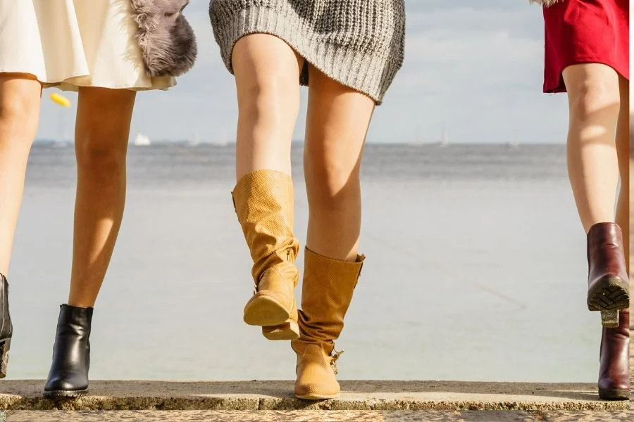 Three women's legs on a seafront wall, one pair in camel slouch boots, the others in black and burgundy ankle boots