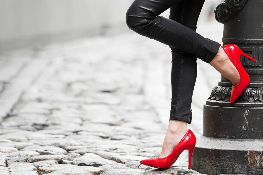 Woman in black leather trousers leaning on a cast-iron lamp post in red stiletto court shoes on cobblestones