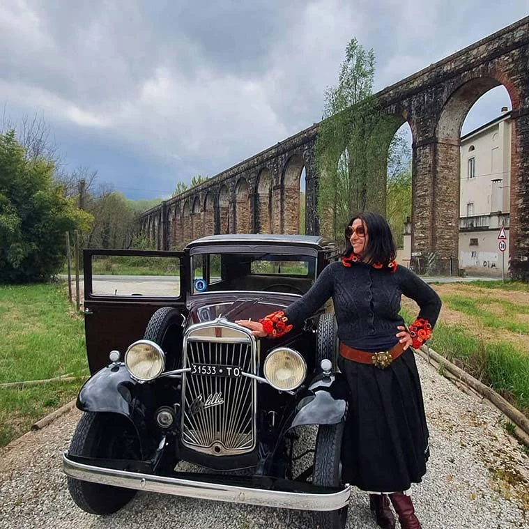 Gracie Opulanza in a black jumper and long skirt posing by a vintage black saloon car, with a stone aqueduct behind