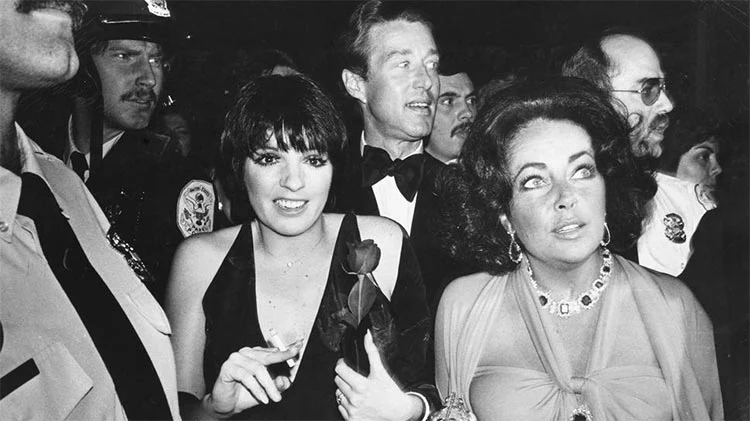 Black-and-white photograph of a woman holding a rose in a dark dress and a woman in a jewelled necklace, in a crowd