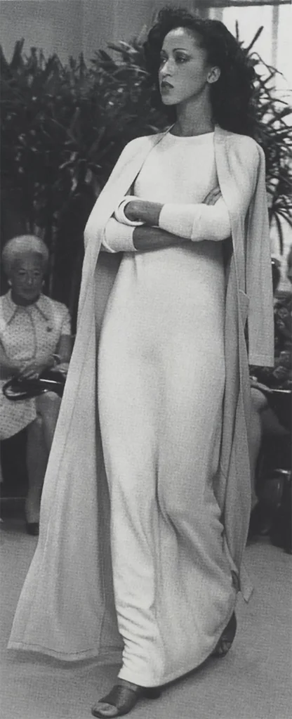 Black-and-white runway photograph of a model in a long white column dress and matching long coat, a seated woman behind