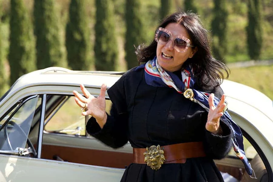 Gracie Opulanza in pink glasses, a navy printed scarf and a brown belt with a gold lion-head buckle, beside a white car