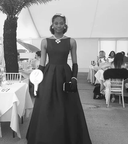 Black-and-white photograph of a woman in a long black sleeveless gown, black gloves and a pearl-drop necklace, in a marquee