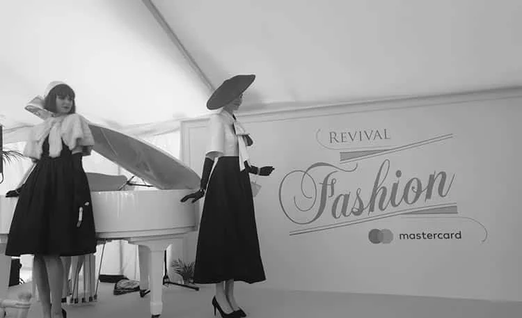 Two models in black full skirts and white jackets by a white grand piano, before a Revival Fashion sign, in black and white