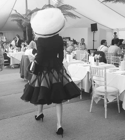 Black-and-white view from behind of a woman in a wide white hat, fitted black jacket and full skirt at Goodwood Revival
