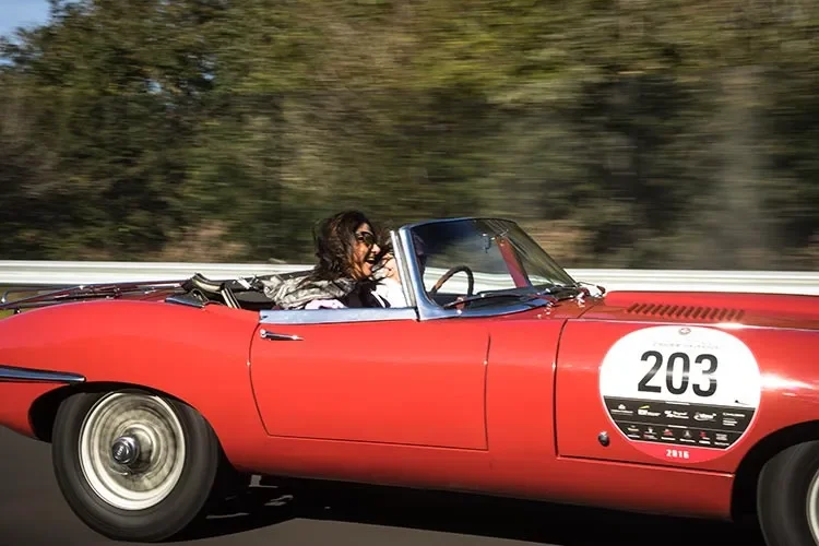 Gracie Opulanza in a red vintage Jaguar open-top car, race number 203, driving past blurred trees