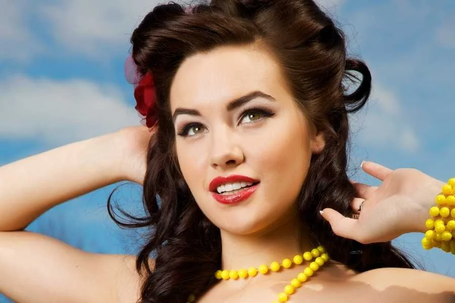 Woman with dark pinned-up curls, a red flower in her hair and a yellow bead necklace, against a blue sky