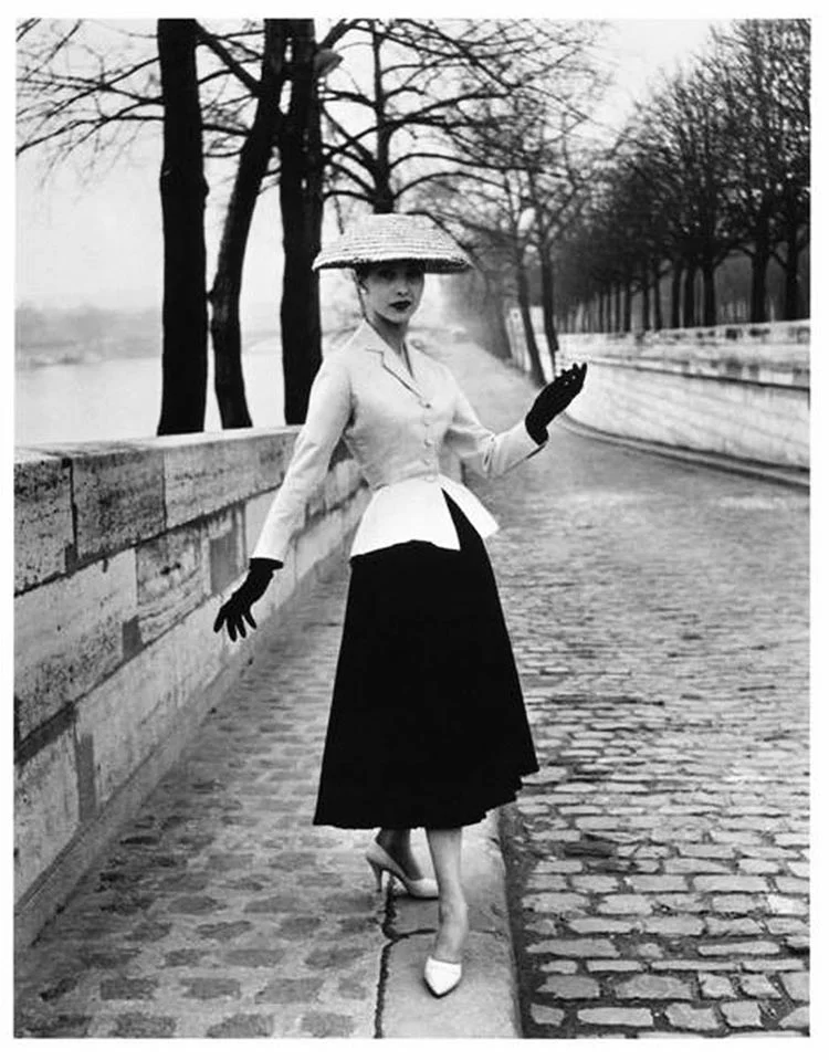 Black-and-white 1947 New Look suit: fitted pale jacket, full black skirt and wide straw hat on a cobbled riverside walk