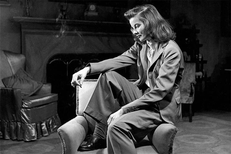 Katharine Hepburn seated in an armchair in a tailored trouser suit, black-and-white photo