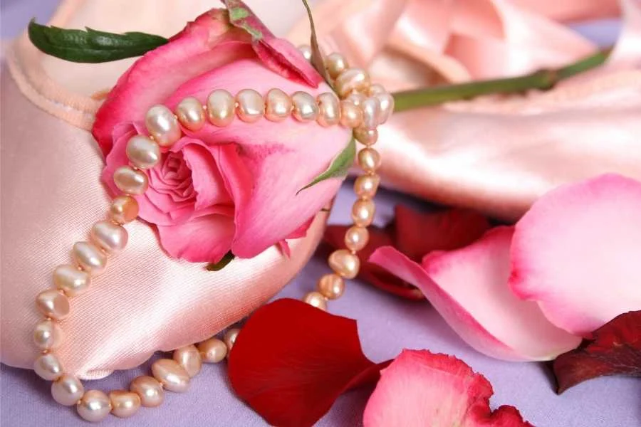 A string of pearls laid over a pink rose and satin ballet shoes, with scattered petals