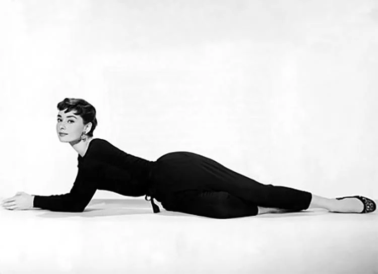 Black-and-white studio photograph of Audrey Hepburn lying on the floor in a black top, trousers and flat shoes