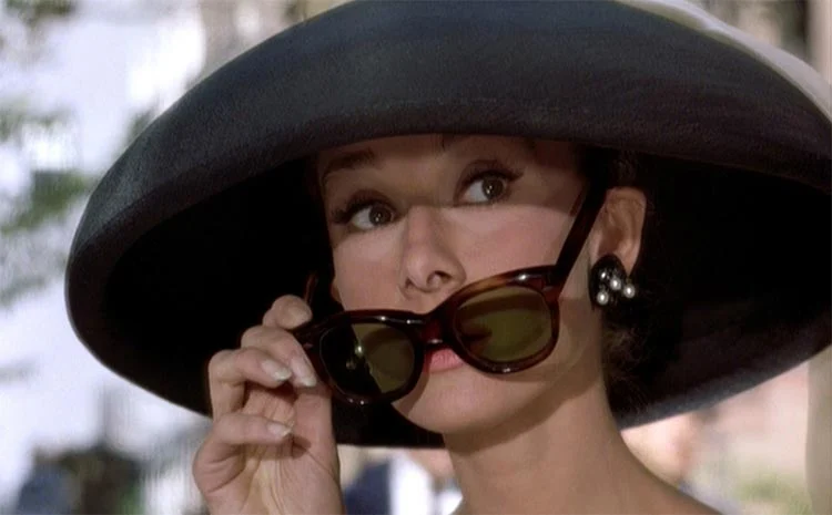 Audrey Hepburn in a wide-brimmed black hat, lowering her tortoiseshell sunglasses with one hand
