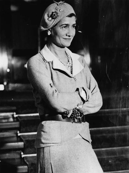 Black-and-white photograph of Coco Chanel in a beret with a flower brooch, a tweed suit and long pearls