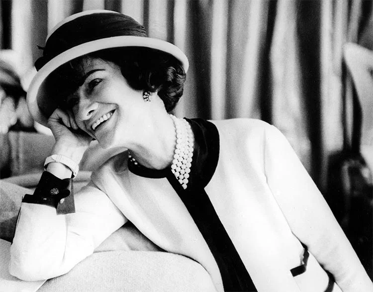 Black-and-white photograph of Coco Chanel smiling in a brimmed hat, pearl necklace and a cream jacket with black trim