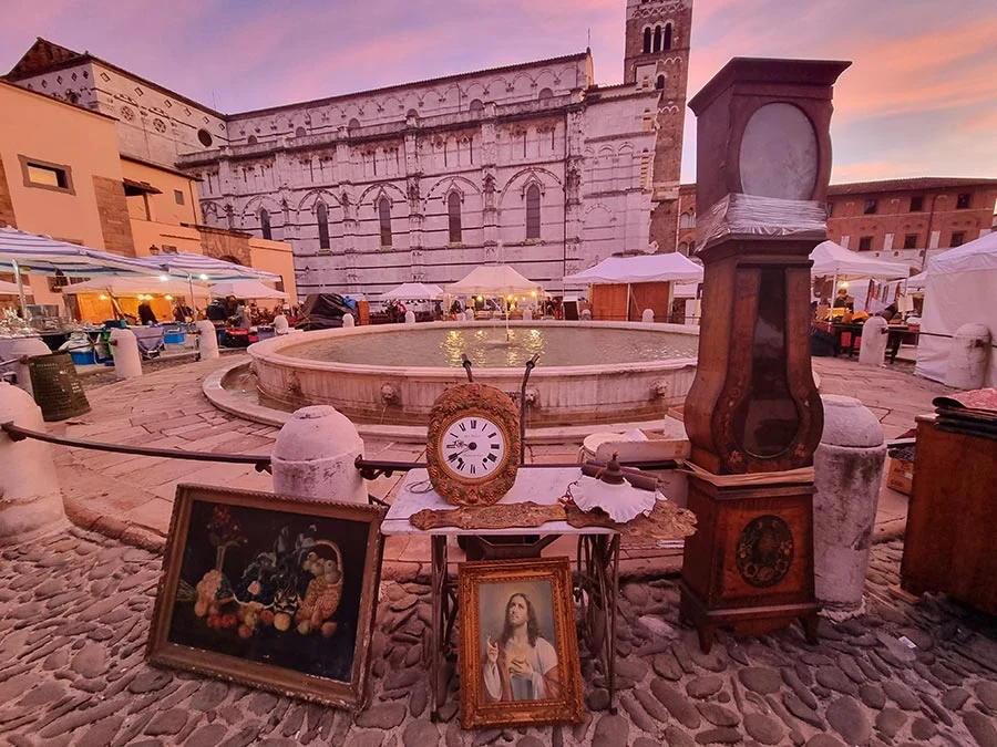 Vintage market in Lucca at dusk: an old clock, framed paintings on the cobbles and a fountain below a marble church
