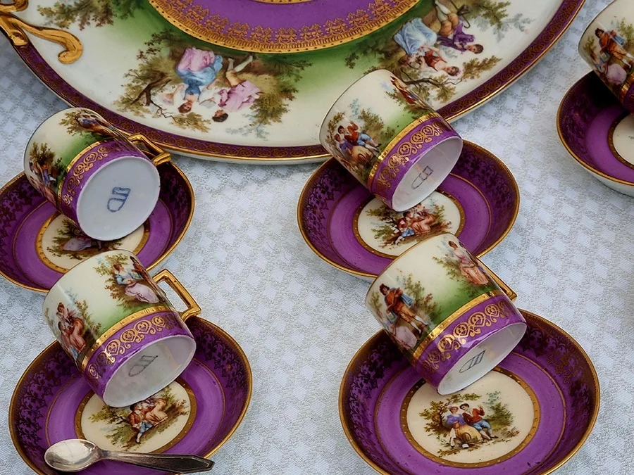 Violet and gold porcelain coffee cups and saucers painted with pastoral scenes, laid out on a white cloth at the market