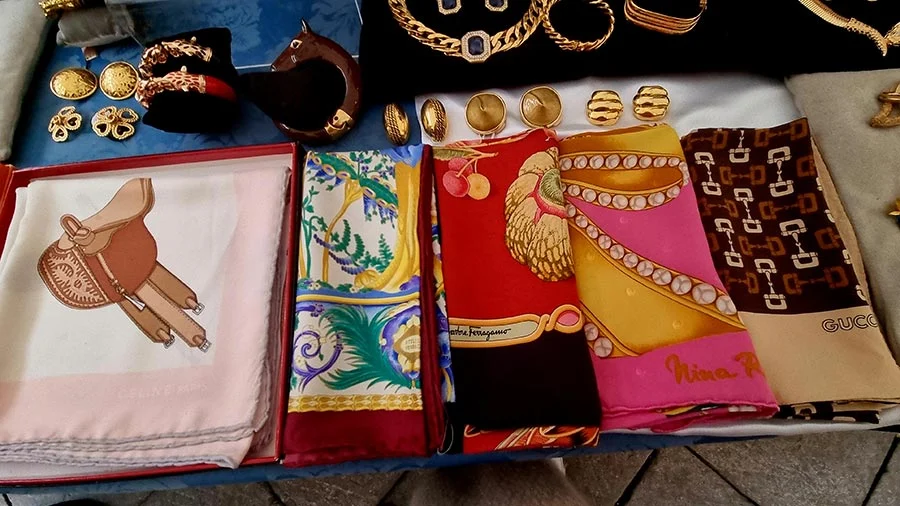 Vintage silk scarves laid out on a market stall, with a saddle print and labels reading Ferragamo, Nina Ricci and Gucci