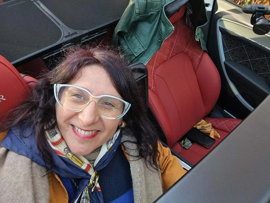 Gracie Opulanza smiling in pale blue-framed glasses and a silk scarf, in the red leather seat of a convertible