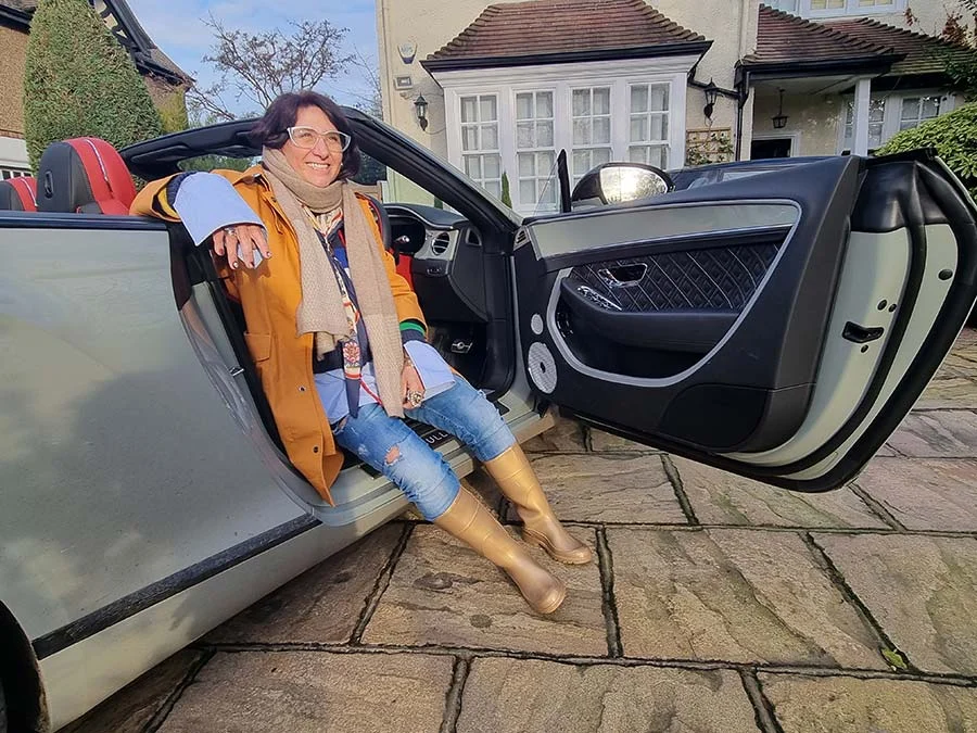 Gracie Opulanza seated in the open door of a pale Bentley convertible, in an ochre jacket, jeans and tan boots
