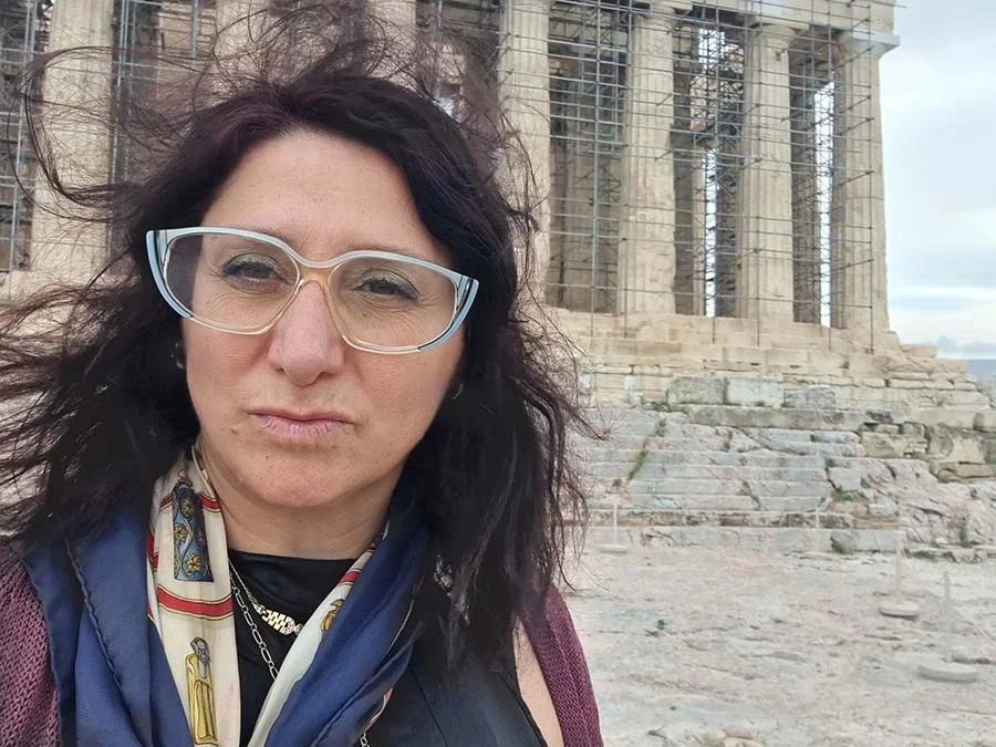 Gracie Opulanza in pale blue-framed glasses and a patterned silk scarf, in front of a columned temple under scaffolding