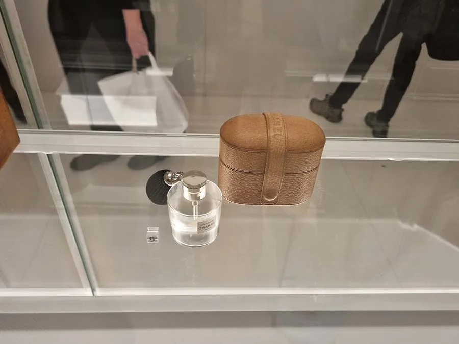 Chanel perfume atomiser beside a tan leather travel case in a museum display case