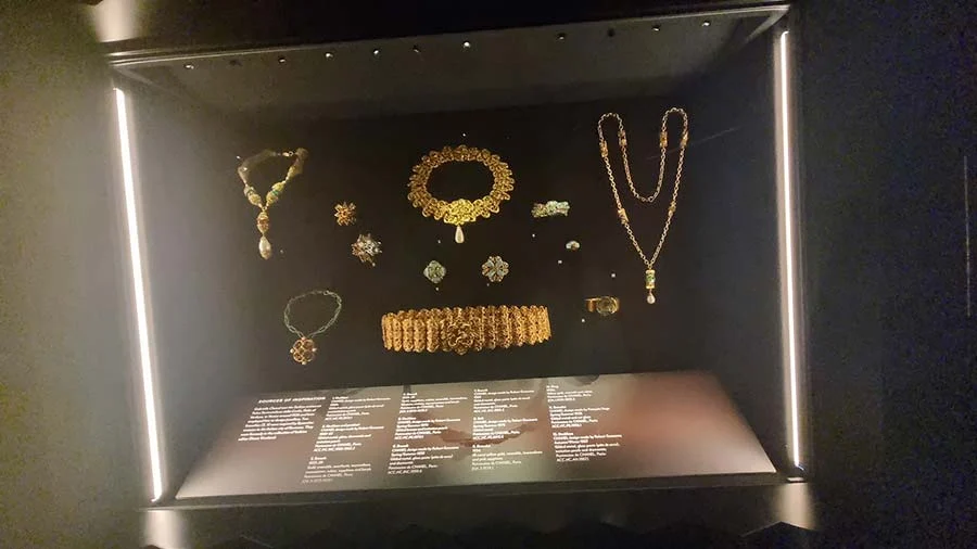 Display case of gold-toned Chanel jewellery in a dark gallery: necklaces, a collar, a bracelet and brooches