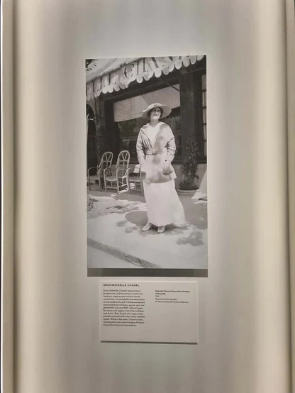 Black-and-white photograph of Mademoiselle Chanel in a hat and long white dress outside a shopfront, on a museum panel