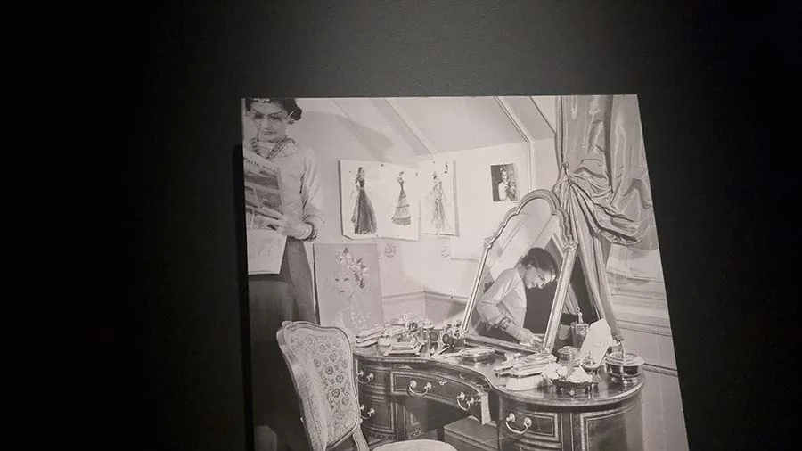 Gallery display of a black-and-white photograph of a woman beside a mirrored dressing table and fashion sketches