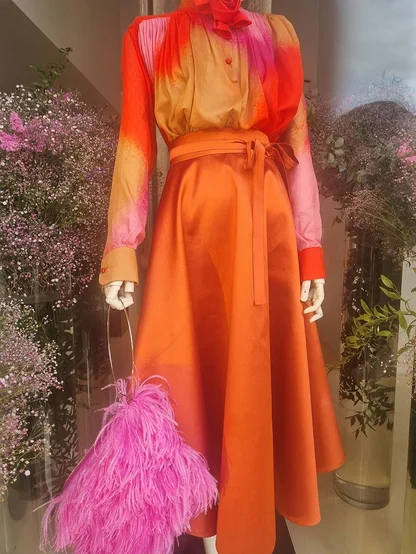 Shop display of a printed orange and pink blouse, a full orange satin skirt and a pink feather bag