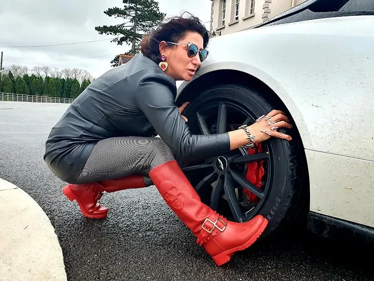 Gracie Opulanza crouching by the wheel of a white Aston Martin DB11 in a leather jacket and red buckled boots