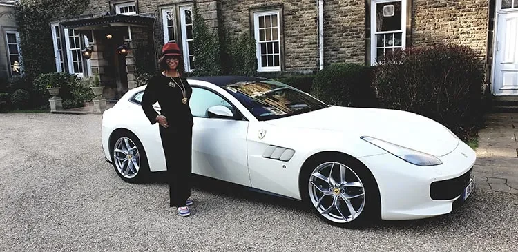 Gracie Opulanza in black and a red hat beside a white Ferrari GTC4Lusso T outside a stone house
