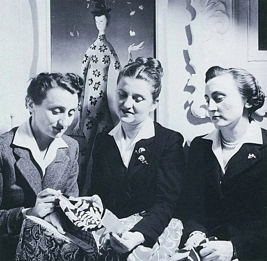 Vintage black-and-white photograph of the Fontana sisters in dark jackets with white collars, one holding a printed fabric