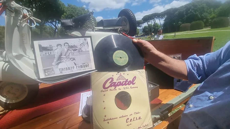 A hand holding a Capitol record and its sleeve beside the Vespa and the framed Roman Holiday still