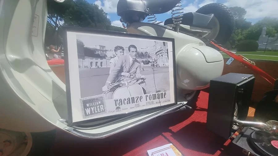 Framed black-and-white lobby card for Vacanze romane, two people on a Vespa, propped against a cream vintage scooter