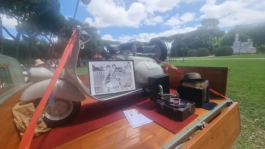 Cream Vespa on a wooden trailer with a framed film still, a gramophone record and a hat, in a Rome park