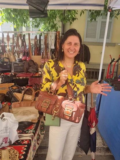 A smiling woman in a yellow printed top holding a brand new tan leather Coach bag with gold buckles at a bag stall