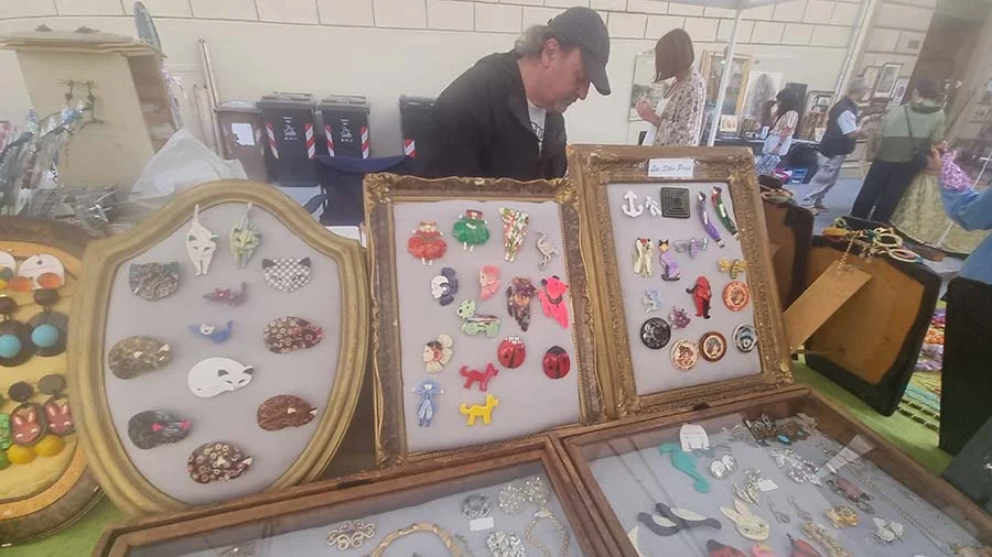 A man in a cap browsing gilt-framed trays of colourful enamel animal brooches on a stall