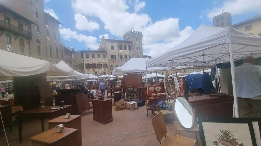 White market canopies across a piazza, with old furniture, a mirror and framed prints on sale beneath a stone tower