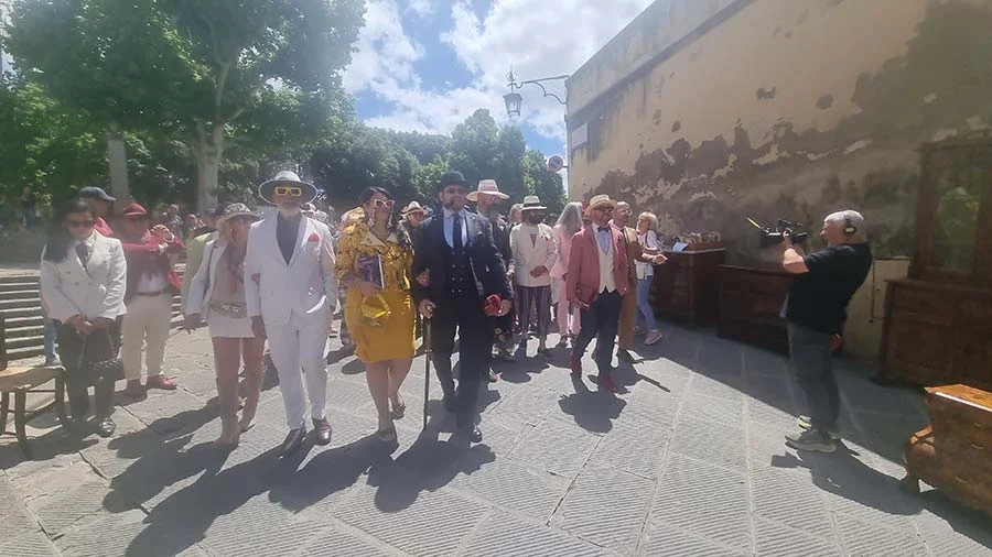 A group in vintage-style suits and dresses walking down a paved street in Arezzo while a man films them